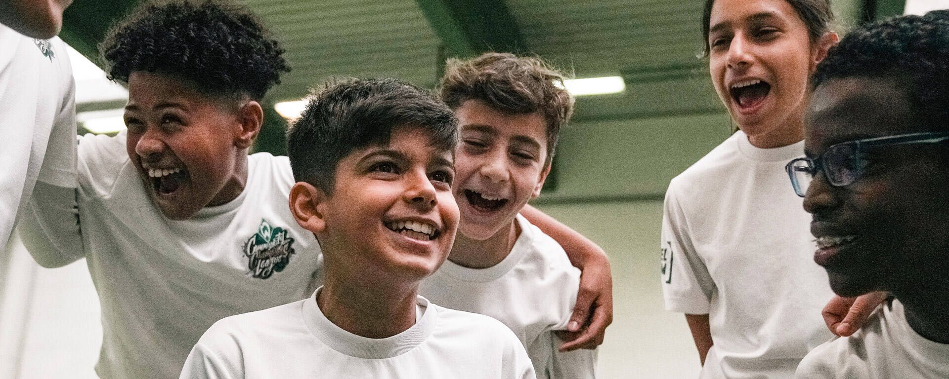 Eine Gruppe lächelnder Kinder in weißen Hemden, die dicht beieinander im Innenraum stehen und fröhlich und interessiert wirken.