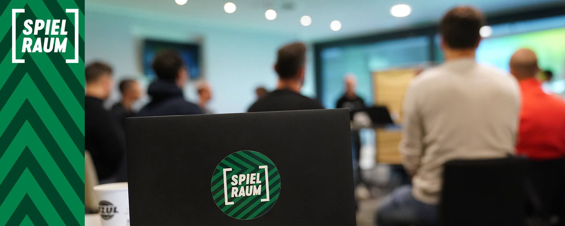 Laptop mit „SPIELRAUM“-Aufkleber im Fokus, umgeben von einer Gruppe von Menschen in einem Besprechungsraum mit unscharfem Hintergrund.