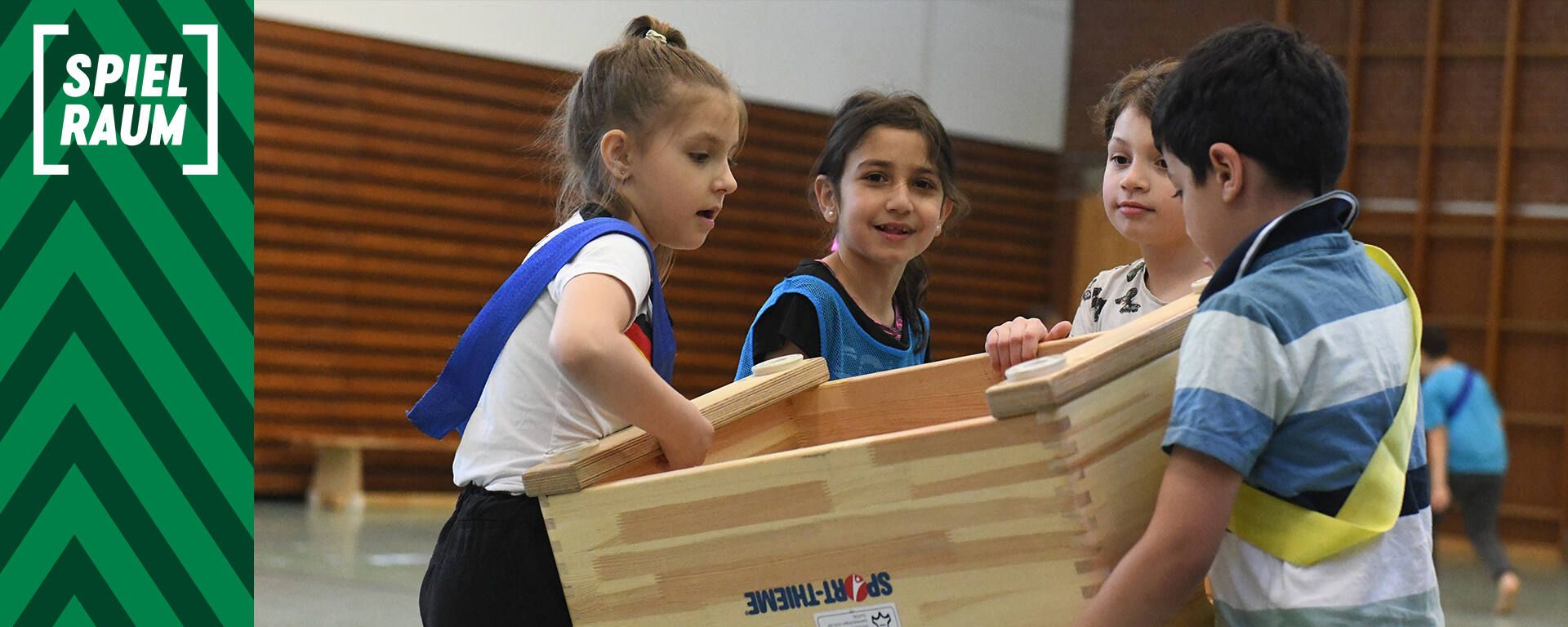 Kinder spielen in einer Turnhalle und heben gemeinsam eine Holzkiste. Sie tragen bunte Sporttrikots. Links ist ein Schild mit der Aufschrift „Spielraum“ zu sehen.