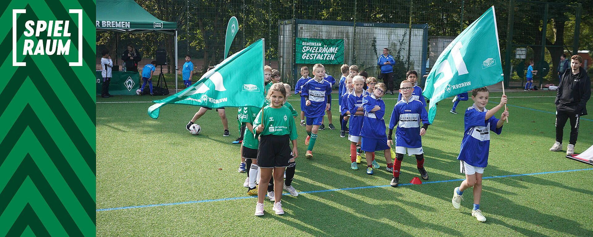 Kinder in Sportuniformen mit grünen Fahnen führen eine Gruppe auf einem Fußballfeld an, links ist ein Banner mit der Aufschrift „SPIELRAUM“ zu sehen.