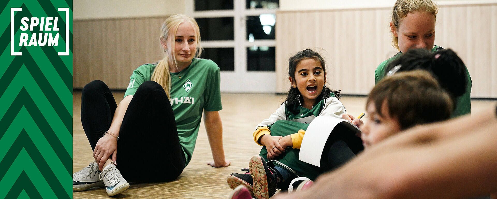 Kinder und junge Erwachsene sitzen auf dem Boden einer Turnhalle und unterhalten sich. Links ist ein grünes Banner mit der Aufschrift „SPIELRAUM“ zu sehen.