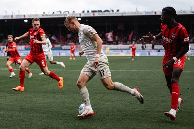 Marco Grüll in weißem Trikot dribbelt den Ball während eines Spiels auf einem Rasenfeld an Gegnern in roten Trikots vorbei. Im Hintergrund sind das Stadion und Zuschauer zu sehen.