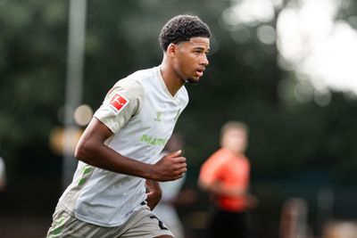 Ein Fußballspieler in weiß-beigem Trikot rennt über einen Rasen, im Hintergrund ist ein verschwommener Schiedsrichter zu sehen.
