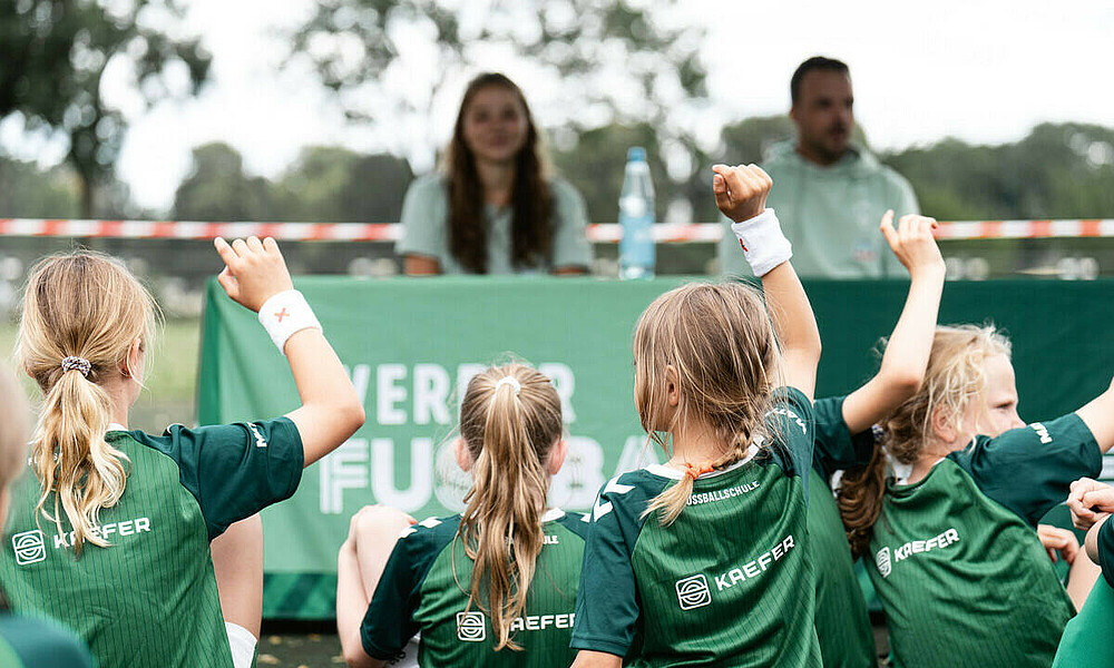 Bundesliga-Spielerin Lina Szaraz und Nachwuchs-Trainer Alex Kluge sitzen im Hintergrund. Im Vordergrund melden sich mehrere Mädchen, um eine Frage zu stellen.