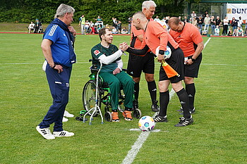 Matthias Hartmann im Rollstuhl im Mittelkreis, klatscht dem Schiedsrichter ab und übergibt den Ball. 