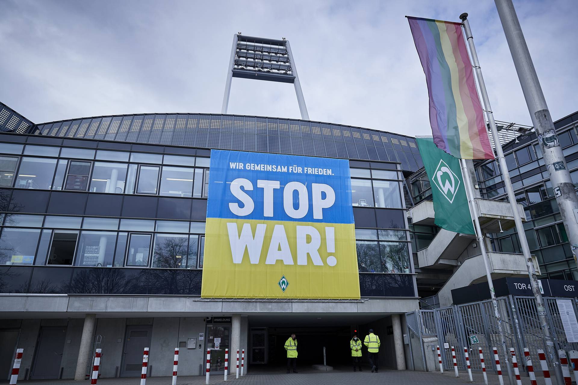 Stadioneingang mit einem großen „Stoppt den Krieg!“-Transparent in Blau und Gelb. In der Nähe wehen zwei Flaggen, eine davon in Regenbogenfarben.