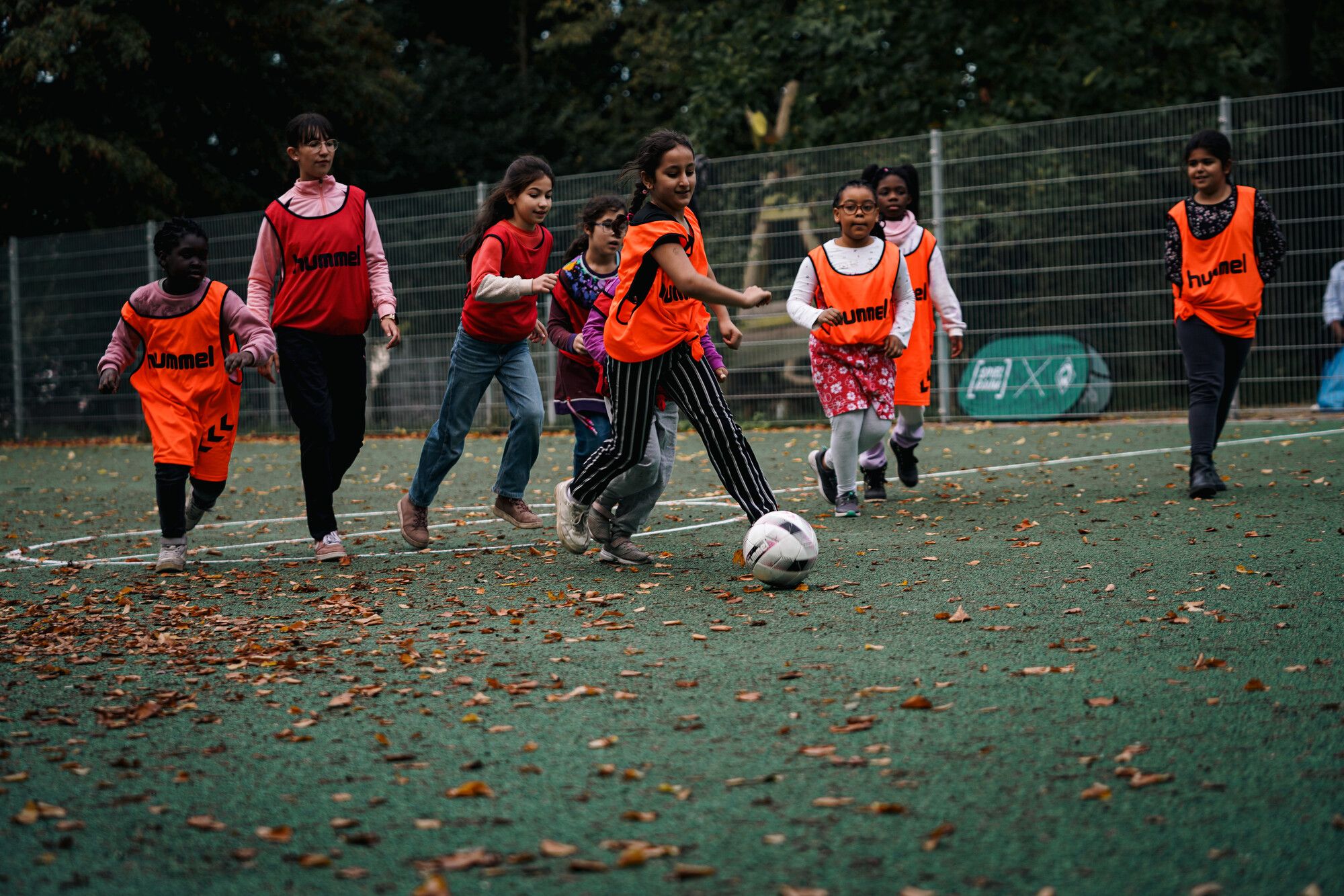 Eine Gruppe von Kindern auf einem Bolzplatz rennt hinter einem Ball hinterher. Sie tragen orangene und rote Leibchen.