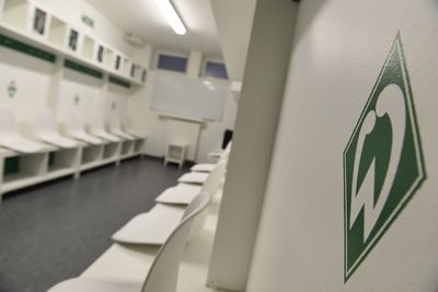 Leerer Umkleideraum mit weißen Stühlen, grünen Logos an den Wänden und einer Weißwandtafel im Hintergrund.