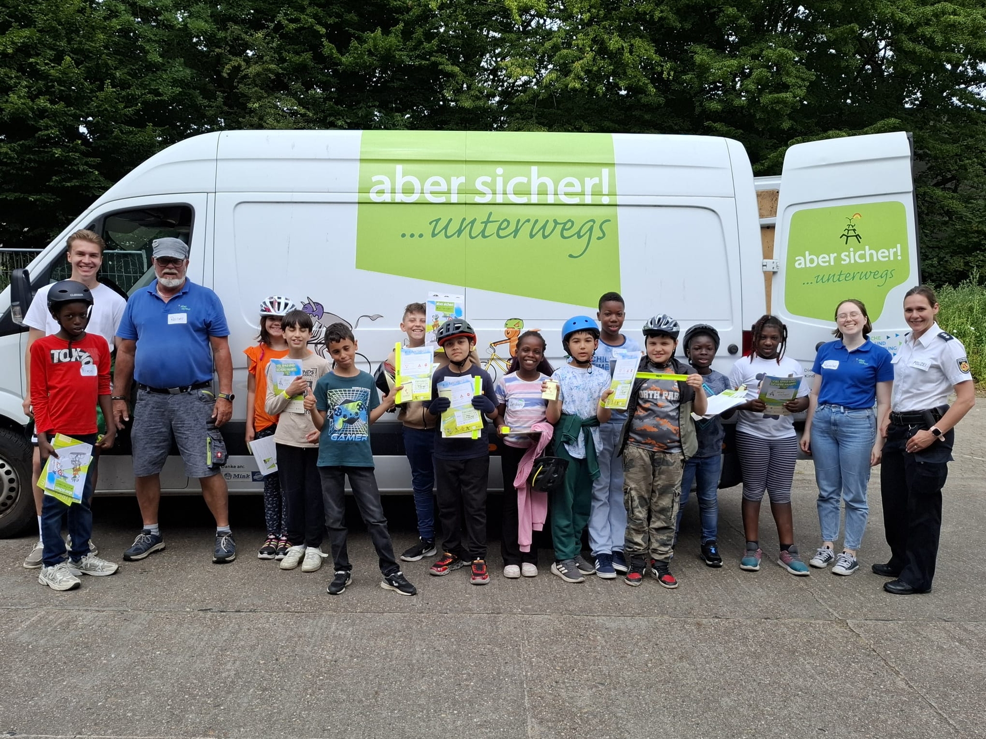 Eine Gruppe von Kindern und Erwachsenen steht vor einem weißen Lieferwagen mit der Aufschrift „aber sicher! ...unterwegs“, hält Karten in den Händen und trägt Helme.