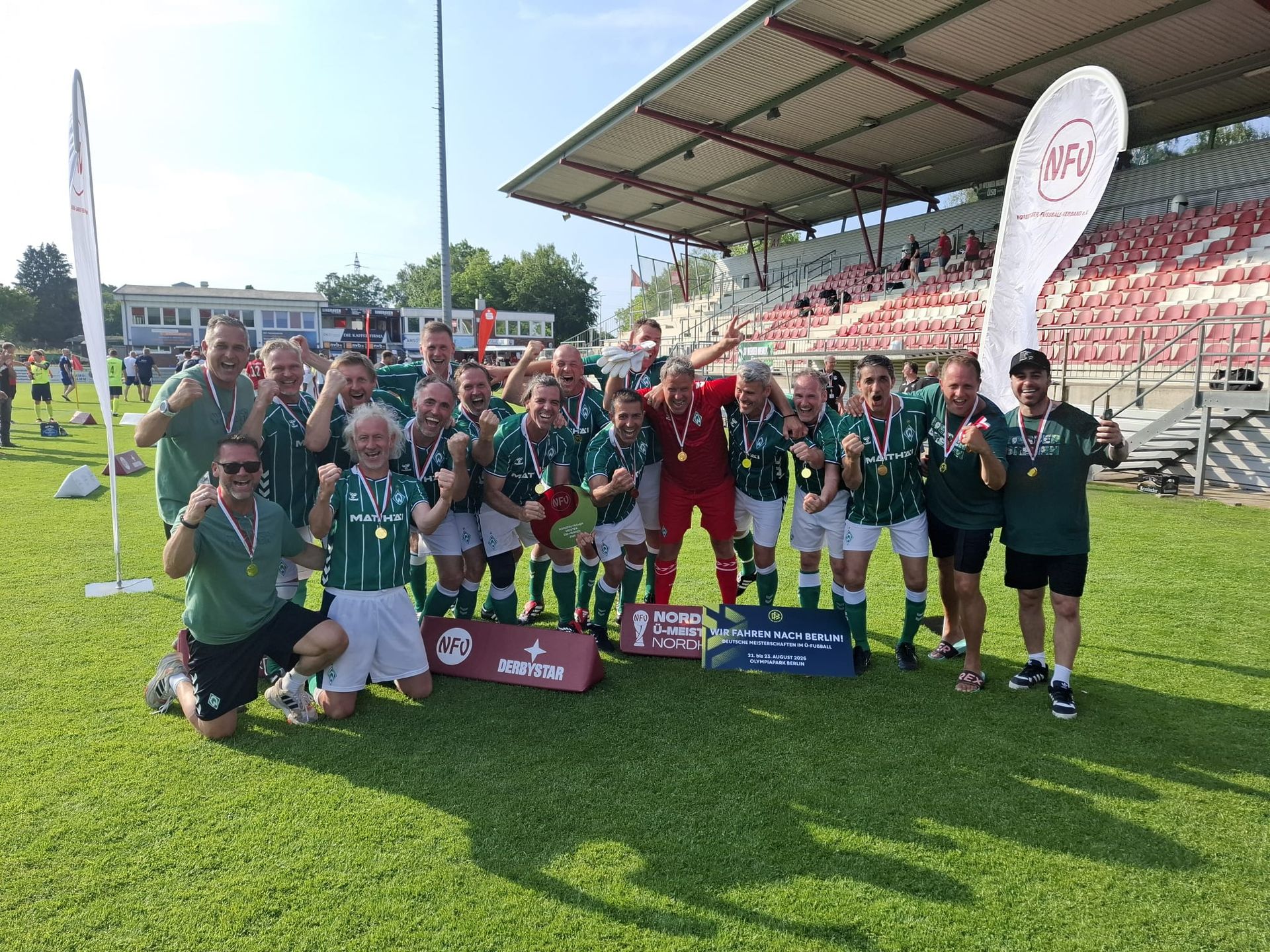 Eine Fußballmannschaft in grünen Trikots feiert mit Medaillen und posiert auf dem Spielfeld vor einem leeren Stadion. Banner und eine Flagge sind zu sehen.