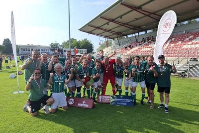 Eine Fußballmannschaft in grünen Trikots feiert mit Medaillen und posiert auf dem Spielfeld vor einem leeren Stadion. Banner und eine Flagge sind zu sehen.