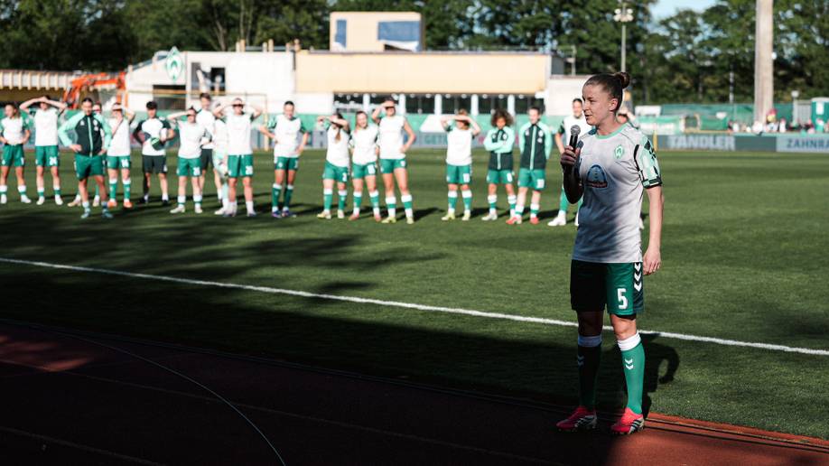 Eine Fußballspielerin steht mit einem Mikrofon auf dem Spielfeld, während ihre Teamkolleginnen in grünen Trikots im Hintergrund aus der Ferne zuschauen.