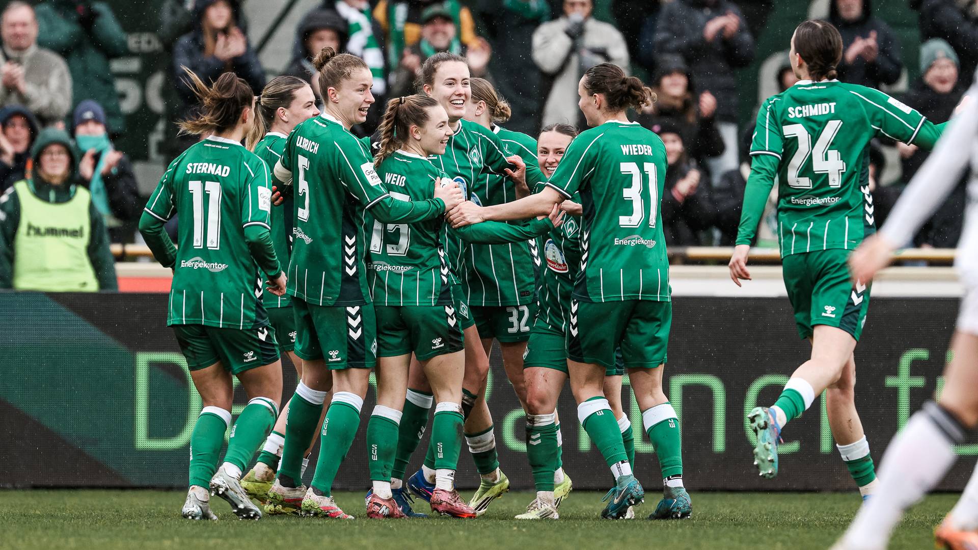 Eine Frauenfußballmannschaft in grünen Trikots feiert gemeinsam auf dem Spielfeld. Zuschauer verfolgen das Geschehen von den Tribünen aus.