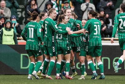 Eine Frauenfußballmannschaft in grünen Trikots feiert gemeinsam auf dem Spielfeld. Zuschauer verfolgen das Geschehen von den Tribünen aus.