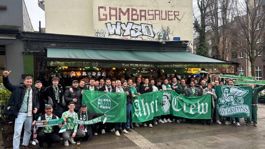Eine Gruppe begeisterter Fußballfans, die grüne Fahnen und Schals in den Händen halten und vor einem Pub posieren, über dem Graffiti an der Wand zu sehen ist.