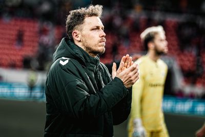 Ein Mann in einer dunklen Jacke klatscht, während er in einem Stadion steht. Im Hintergrund ist eine weitere Person in gelber Kleidung verschwommen zu sehen.