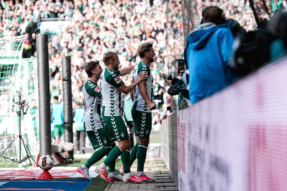 Drei Fußballspieler in grün-weißer Kleidung feiern in der Nähe der Tribüne ein Tor, im Hintergrund ist ein vollbesetztes Stadion zu sehen.