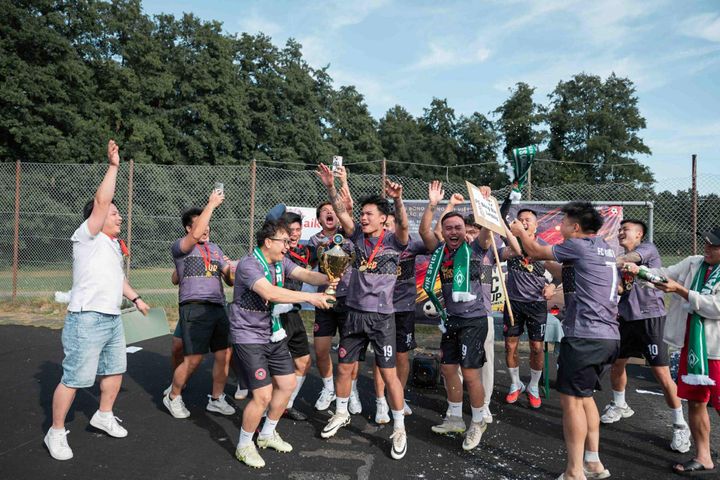 Eine jubelnde Fußballmannschaft feiert mit einem Pokal, trägt Medaillen und bunte Schals an einem sonnigen Tag in der Nähe eines umzäunten Sportplatzes.