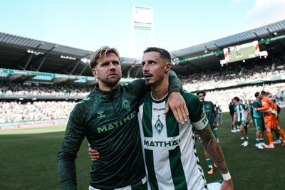 Zwei Fußballspieler in Trikots von Werder Bremen laufen nach einem Spiel in einem vollbesetzten Stadion sichtlich bewegt und mit einem Arm umeinander gelegt über den Rasen.