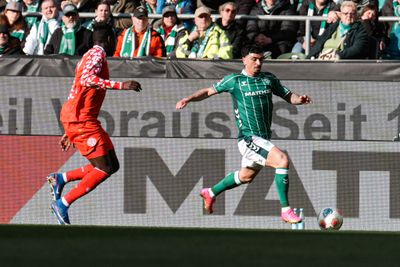 Zwei Fußballspieler auf dem Spielfeld, einer in einem grün gestreiften Trikot und der andere in einem rot gemusterten Trikot, im Hintergrund sind Zuschauer zu sehen.