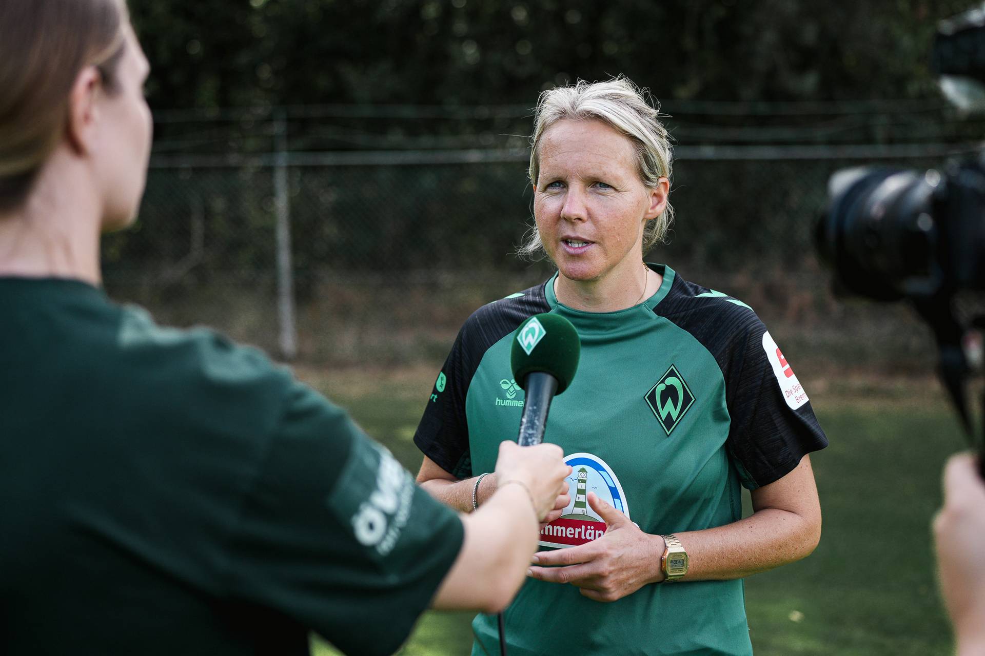 Eine Frau in einem grünen Sporttrikot wird im Freien von einer Person interviewt, die ein Mikrofon mit einem Logo in der Hand hält.