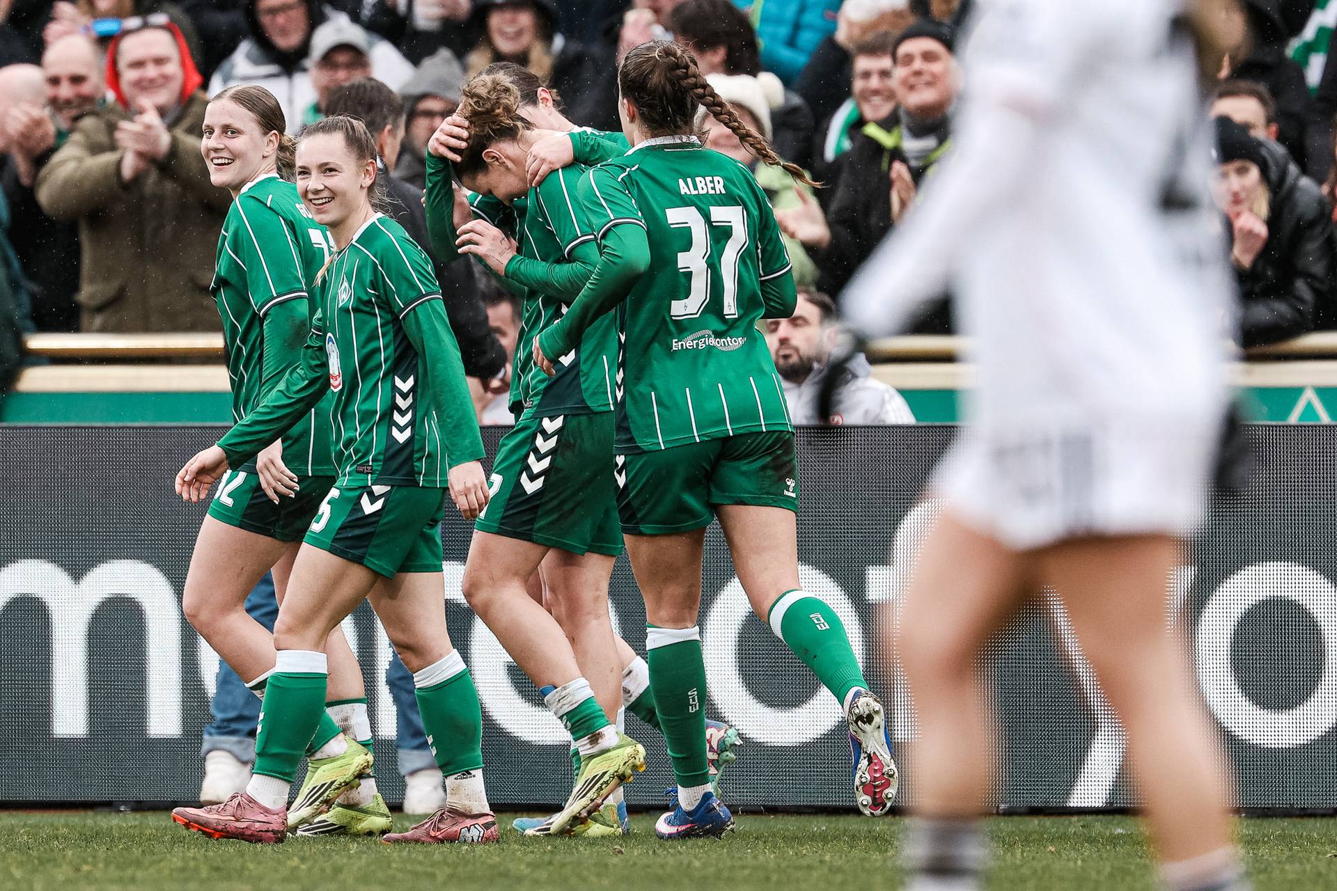 Fußballspieler in grünen Trikots feiern auf dem Spielfeld, während im Hintergrund die Zuschauer jubeln. Im Vordergrund steht ein Spieler in Weiß.