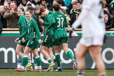 Fußballspieler in grünen Trikots feiern auf dem Spielfeld, während im Hintergrund die Zuschauer jubeln. Im Vordergrund steht ein Spieler in Weiß.