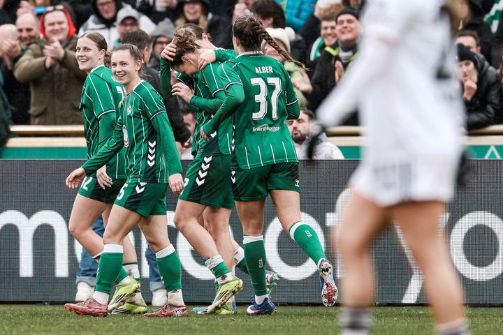 Fußballspieler in grünen Trikots feiern auf dem Spielfeld, während im Hintergrund die Zuschauer jubeln. Im Vordergrund steht ein Spieler in Weiß.