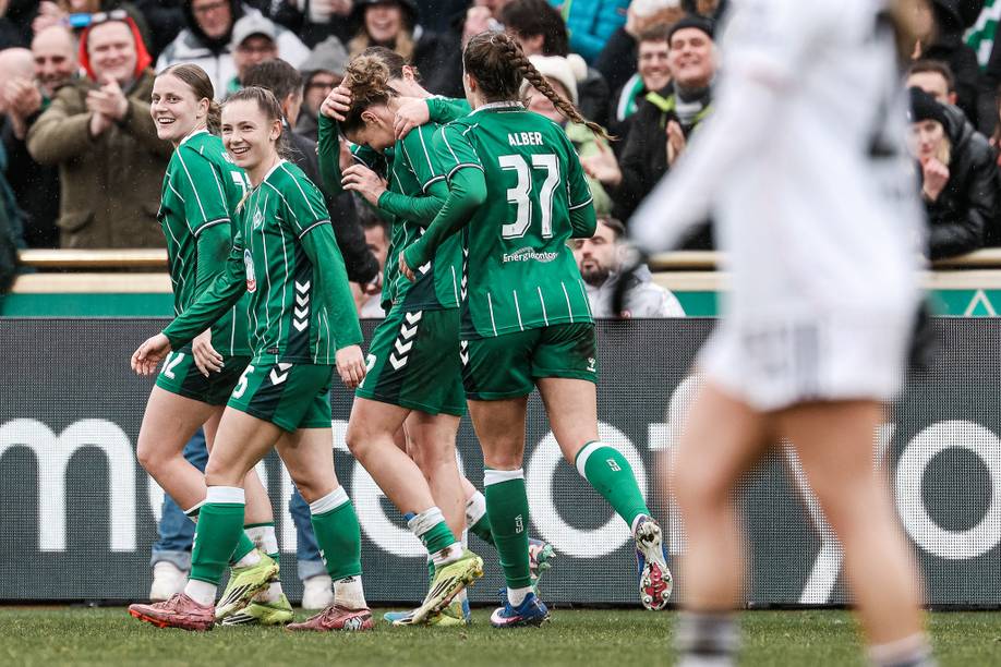 Fußballspieler in grünen Trikots feiern auf dem Spielfeld, während im Hintergrund die Zuschauer jubeln. Im Vordergrund steht ein Spieler in Weiß.