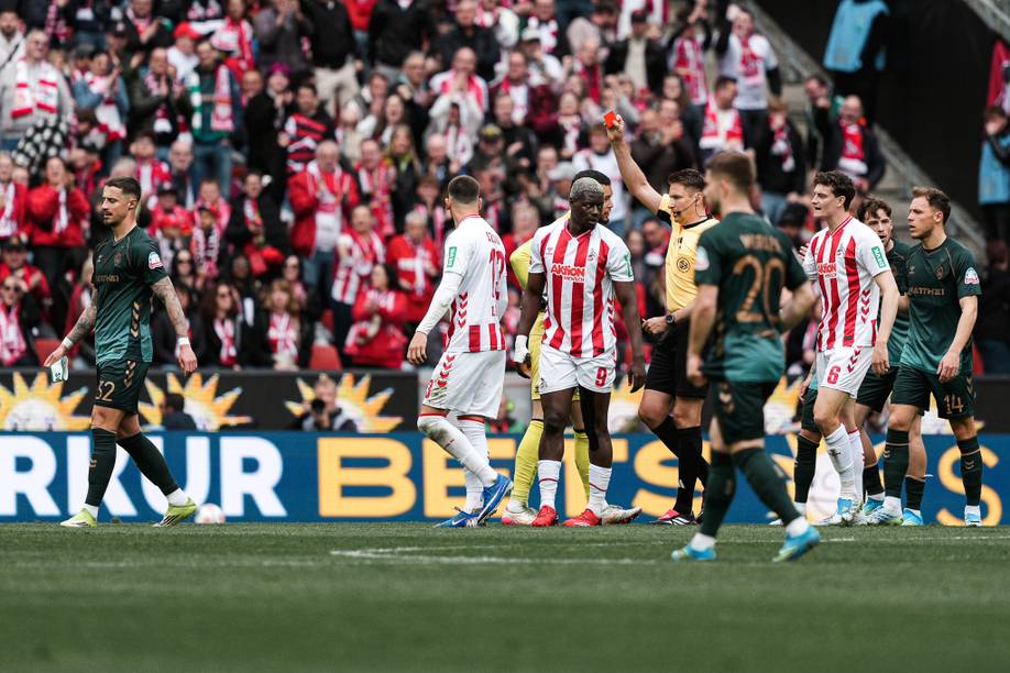 Ein Fußballschiedsrichter zeigt einem Spieler in einem weiß-rot gestreiften Trikot während eines Spiels die rote Karte, im Hintergrund sind Spieler und Fans zu sehen.