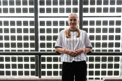 Marie Gmeineder steht in einem gestreiften Shirt vor einem Hintergrund mit Gittermuster und lächelt mit gefalteten Händen in die Kamera.