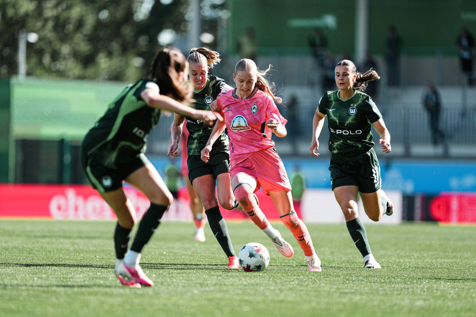 Female soccer player in pink kit dribbles the ball, pursued by three players in green, on a sunny field.
