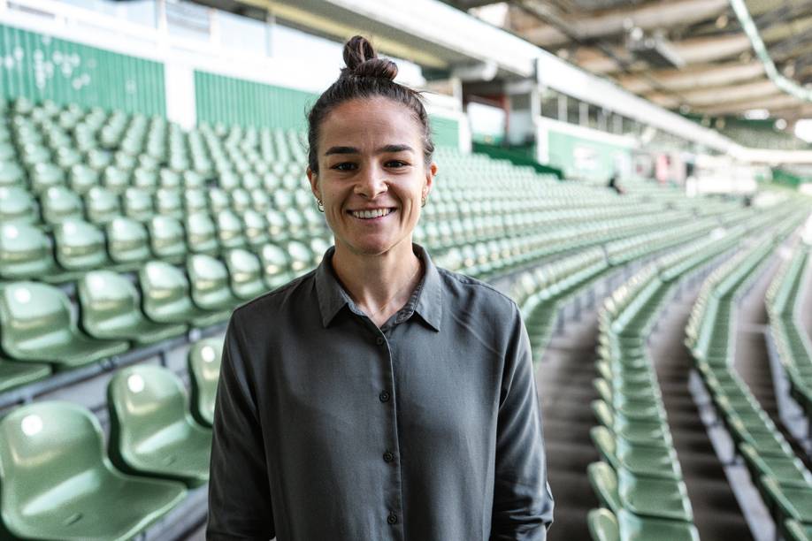 Eine Person mit einem hohen Dutt lächelt in einem leeren Stadion mit Reihen grüner Sitze.