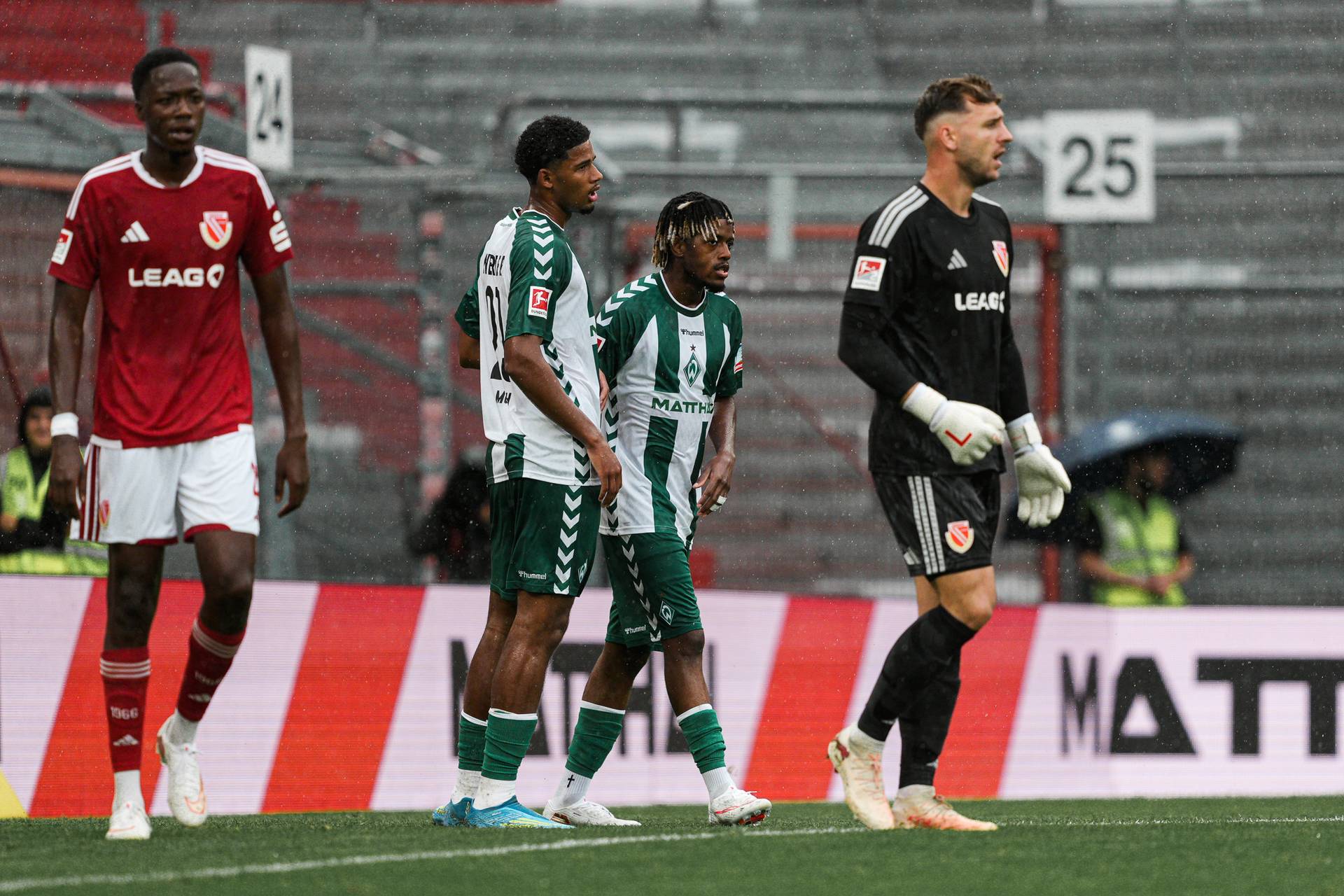 Fußballspieler auf einem Spielfeld während eines Spiels, in roten und grünen Trikots, mit einem Torwart in Schwarz. Im Hintergrund sind die Tribünen des Stadions und Regen zu sehen.