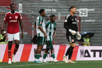 Fußballspieler auf einem Spielfeld während eines Spiels, in roten und grünen Trikots, mit einem Torwart in Schwarz. Im Hintergrund sind die Tribünen des Stadions und Regen zu sehen.