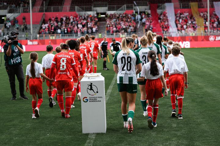 Vor dem Spiel der Google Pixel Frauen-Bundesliga betreten Frauenfußballmannschaften und Kinder-Maskottchen das Spielfeld, während ein Kameramann diesen Moment festhält.