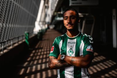 Ein Mann in einem grün-weiß gestreiften Fußballtrikot steht selbstbewusst mit verschränkten Armen in einem sonnendurchfluteten Gang.