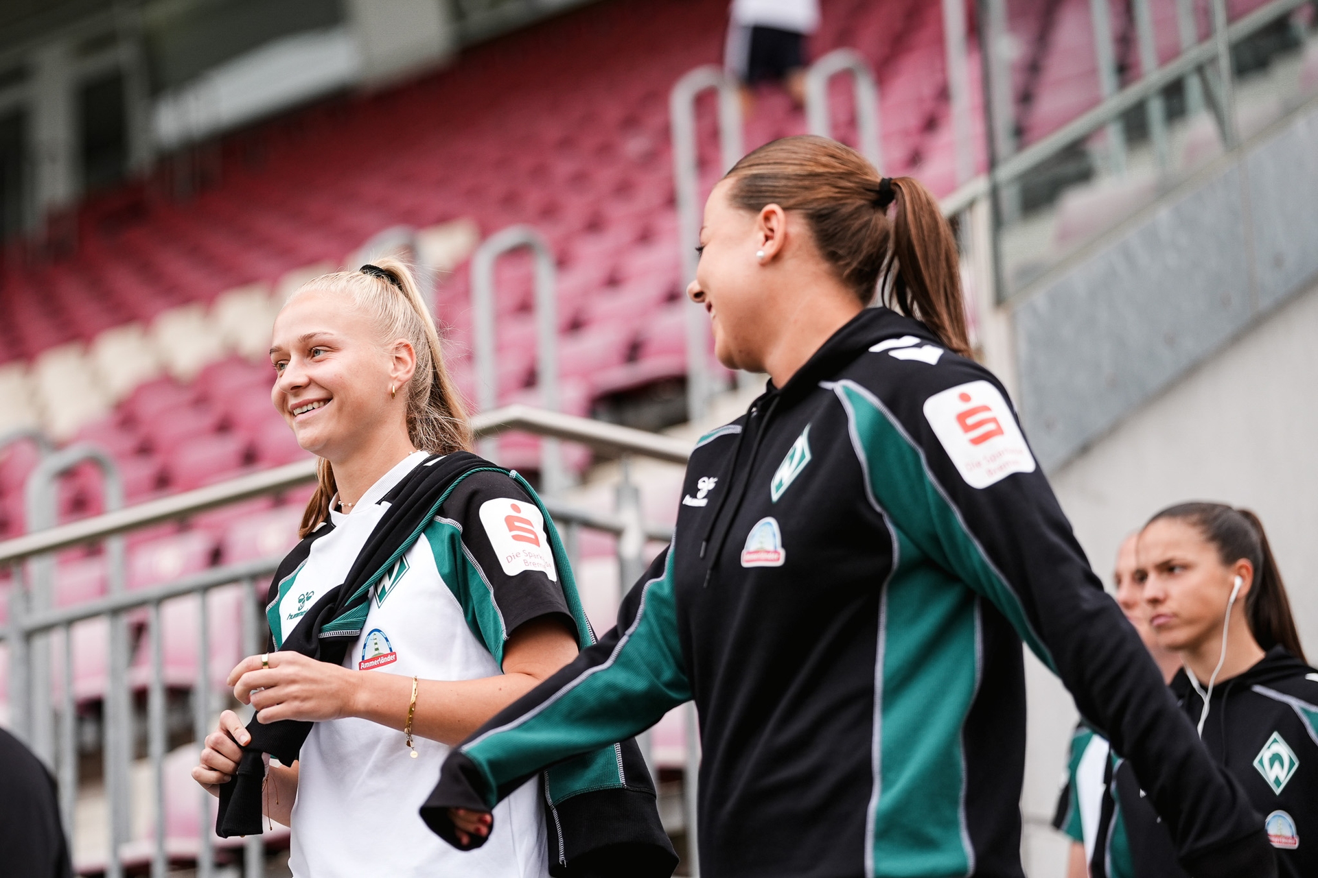 Zwei Frauen in Sportkleidung, die lächelnd durch ein Stadion mit roten Sitzplätzen gehen. Eine dritte Person in ähnlicher Kleidung folgt ihnen.