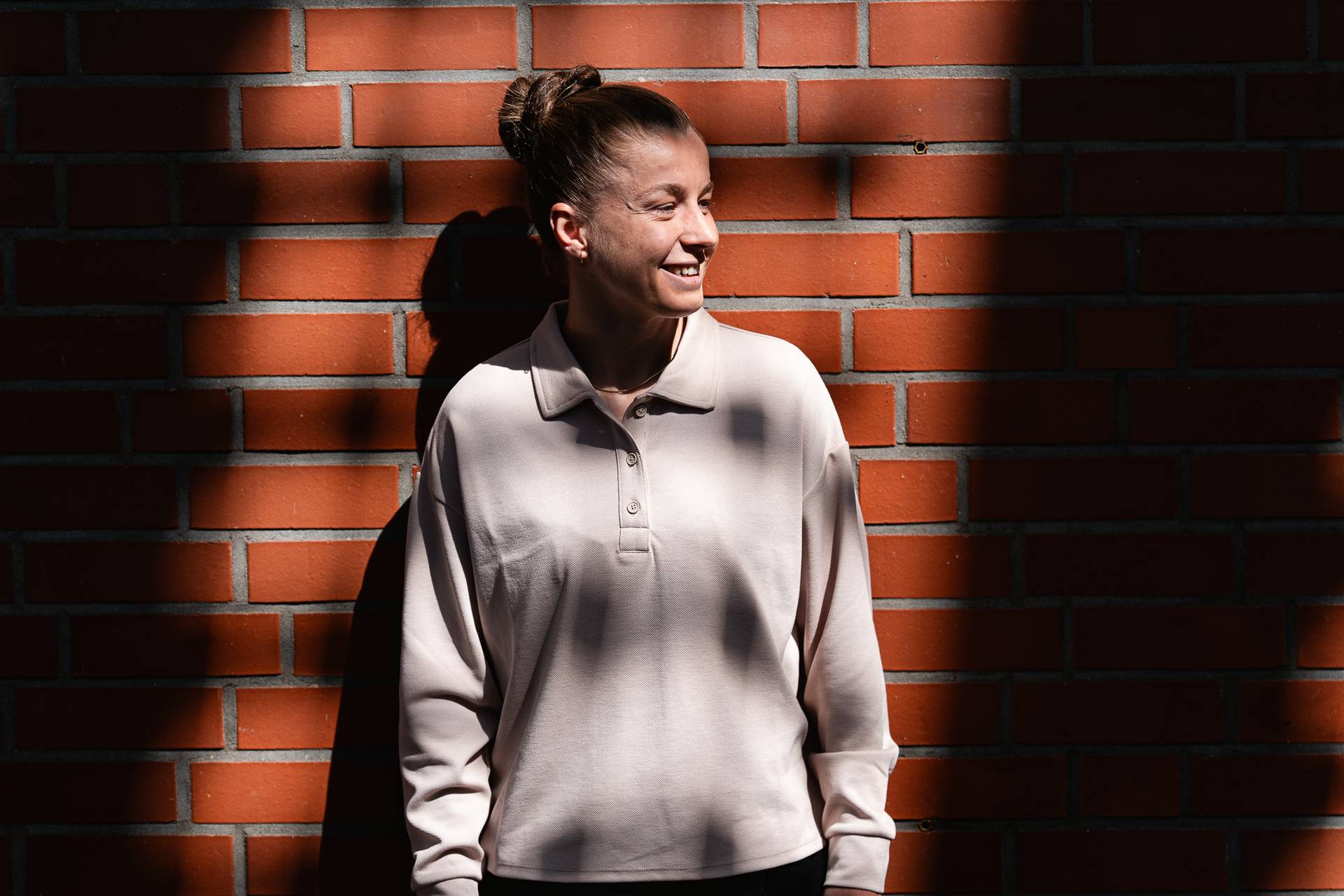 Eine lächelnde Person steht vor einer sonnenbeschienenen Backsteinmauer, trägt ein helles Hemd mit Kragen und hat die Haare hochgesteckt. Schatten zeichnen ein Muster an die Wand.