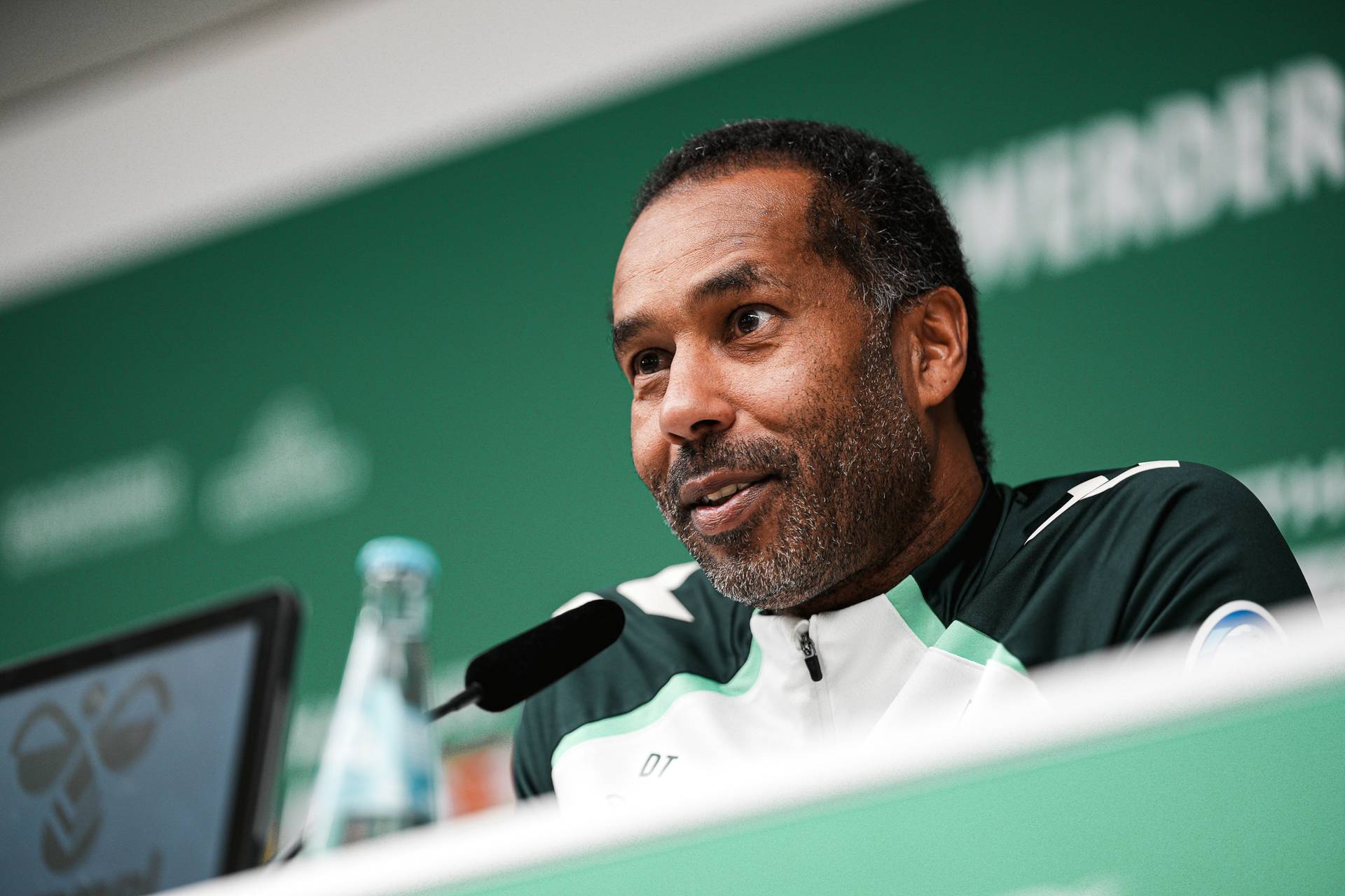 Mann in einer Sportjacke spricht an einem Pressekonferenztisch vor grünem Hintergrund, im Bild ist eine Flasche Wasser zu sehen.