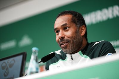 Mann in einer Sportjacke spricht an einem Pressekonferenztisch vor grünem Hintergrund, im Bild ist eine Flasche Wasser zu sehen.