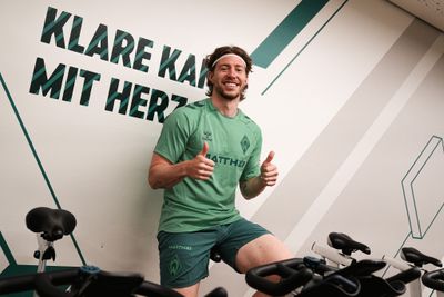 Ein Mann in grüner Sportkleidung sitzt auf einem Heimtrainer in einem Fitnessstudio, hinter ihm ist ein motivierender Wandtext zu sehen; er lächelt und streckt den Daumen nach oben.