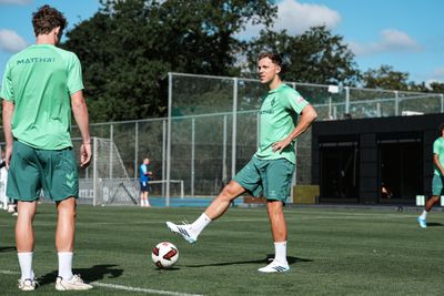 Zwei Fußballspieler in grünen Trainingstrikots stehen auf einem Spielfeld, einer balanciert einen Ball auf seinem Fuß.