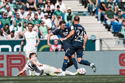 Eine Szene aus einem Fußballspiel, in der ein Spieler in blauer Spielkleidung um den Ball mit einem in Weiß gekleideten Spieler kämpft, der sich mit einem Hechtsprung auf ihn stürzt, während eine lebhafte Zuschauermenge auf den Tribünen zuschaut.