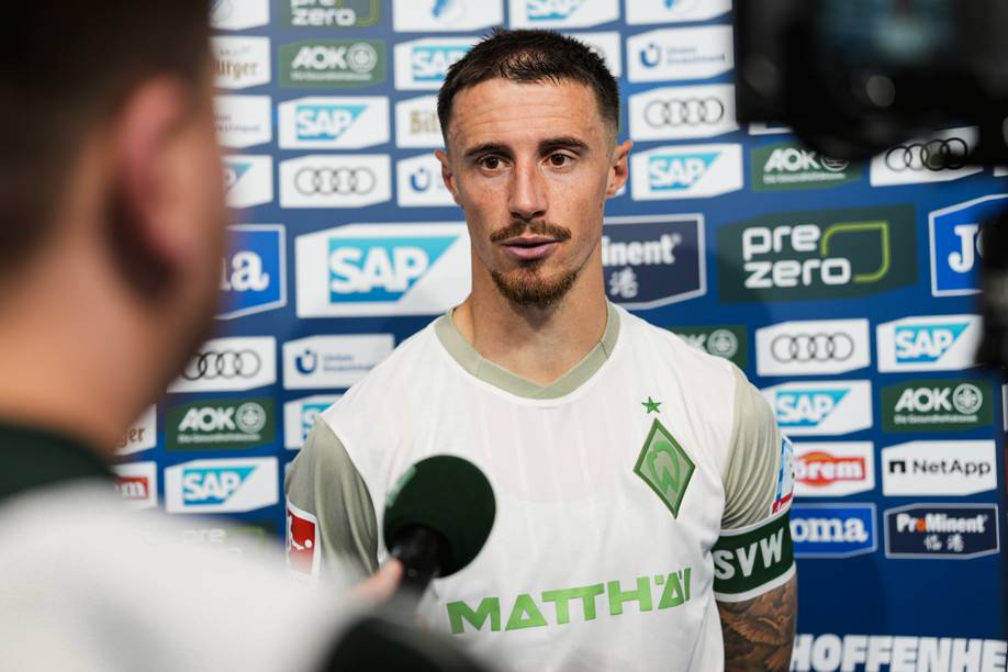 Ein Fußballspieler in einem weißen Trikot wird nach dem Spiel interviewt, wobei die Sponsorenlogos auf blauem Hintergrund zu sehen sind.