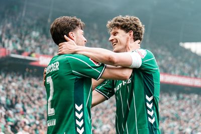 Zwei Fußballspieler in grünen Trikots feiern auf dem Spielfeld, umarmen sich freudig, während im Hintergrund die Zuschauer jubeln.