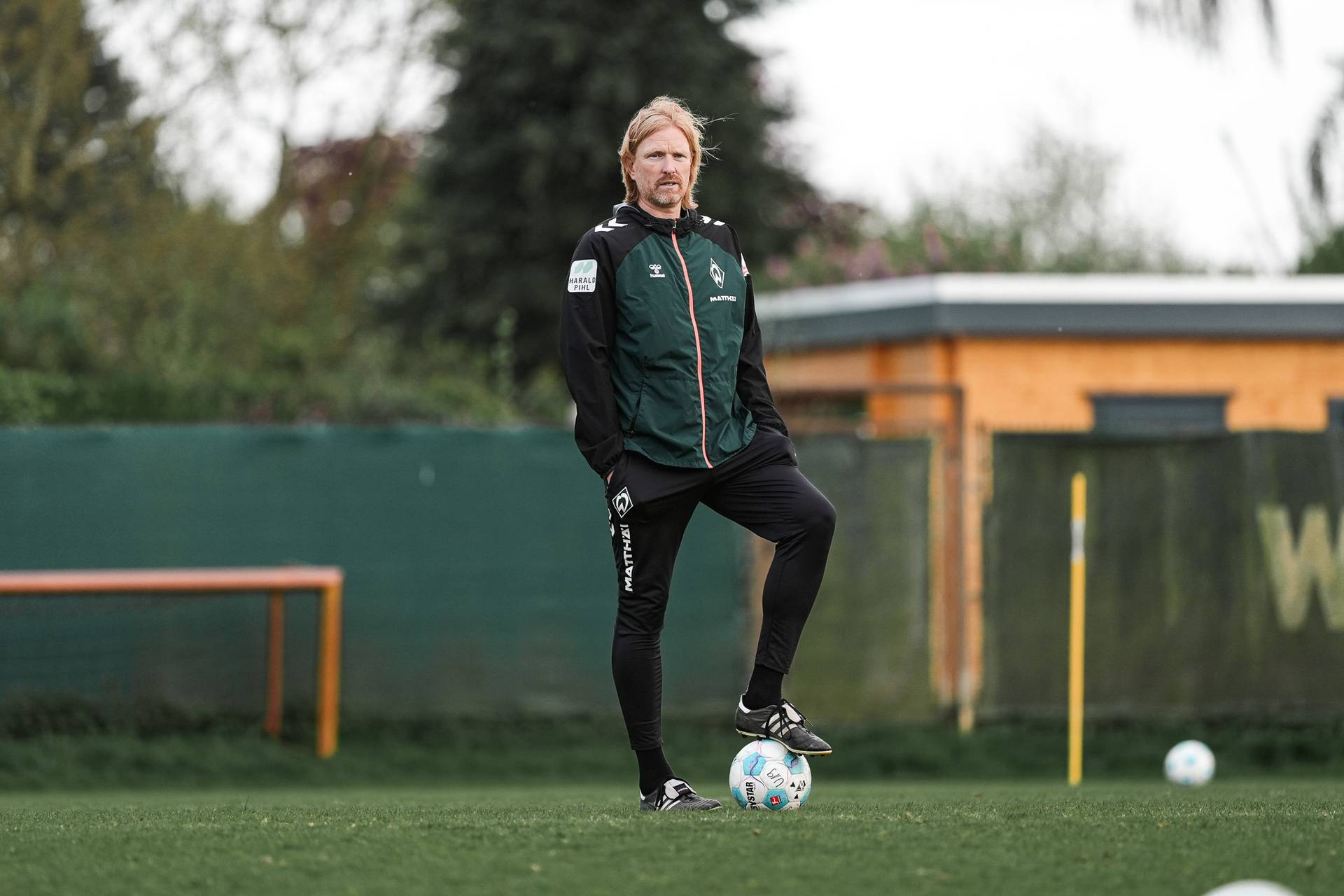 Eine Person in Sportkleidung steht auf einer Wiese, einen Fuß auf einem Fußball abgestützt, im Hintergrund sind ein Gebäude und Bäume zu sehen.