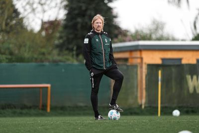 Eine Person in Sportkleidung steht auf einer Wiese, einen Fuß auf einem Fußball abgestützt, im Hintergrund sind ein Gebäude und Bäume zu sehen.
