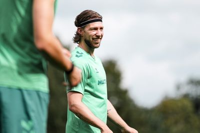 Ein Mann mit langen Haaren und einem Stirnband steht im Freien und lächelt. Er trägt ein hellgrünes Sportshirt, im Hintergrund ist verschwommenes Grün zu sehen.