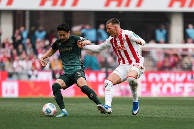 Zwei Fußballspieler, einer in Grün und der andere in rot-weißen Streifen, kämpfen während eines Spiels auf dem Platz um den Ball.